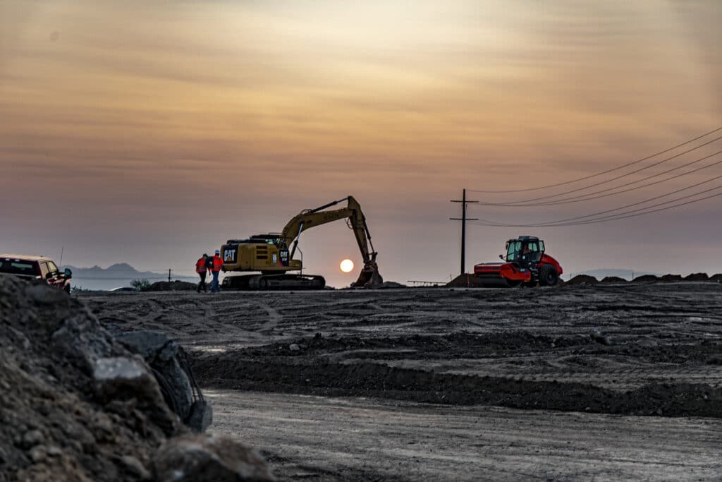 excavator and roller shown in early morning light silhouette on a ridge with operators walking up to start the work day