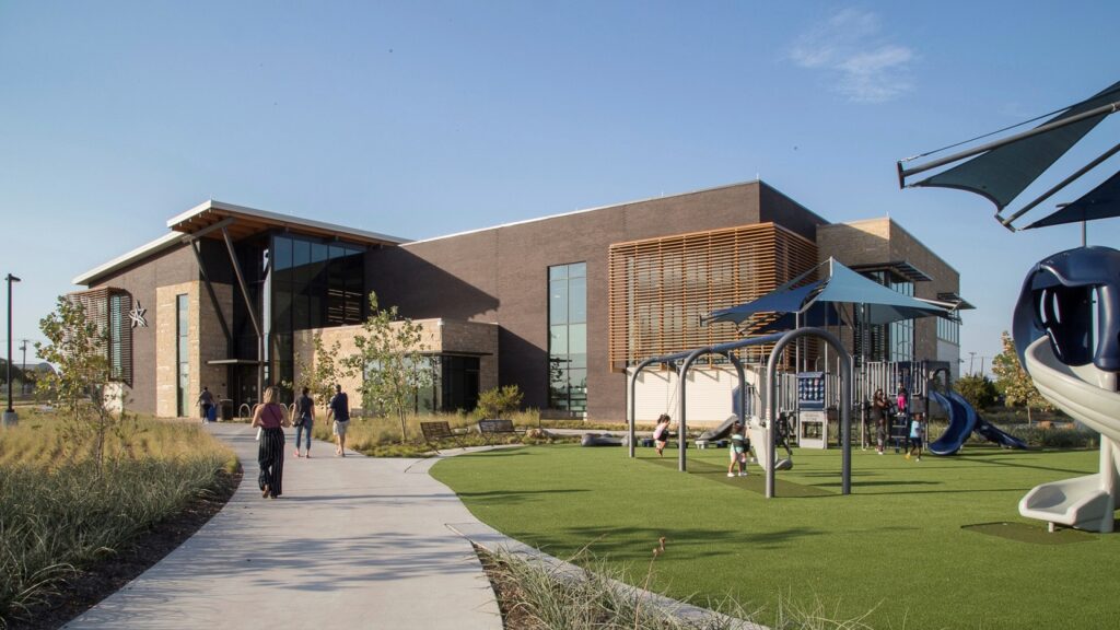 Exterior frontal diurno y entrada principal del Beacon Rec Center en Arlington, Texas, con gente caminando y niños jugando en el frente