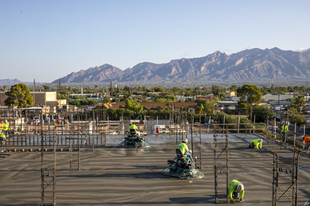 concrete workers use ride-on trowel machines to finish the upper deck of the monastery apartments with the catalina mountains in the background