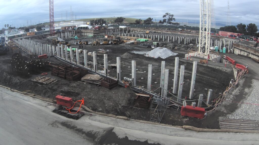 vista diurna del sitio de construcción con filas de pilotes prefabricados y grúas sobre orugas en segundo plano