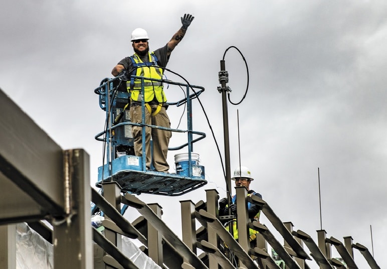 painter standing on man-lift above steel pergola of light rail station waves at camera