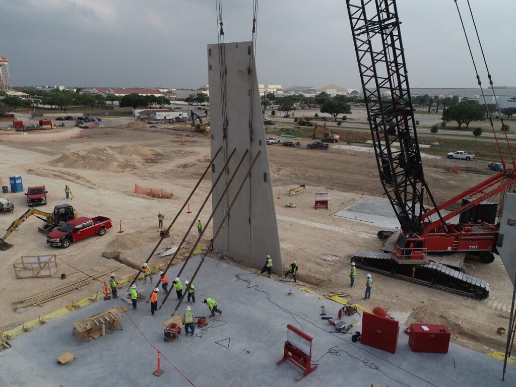 Progress shot of tilt-up construction of Port San Antonio Innovation Center with crawler crane hoisting tilt-up panel and crew of 8 securing support anchors to slab on grade