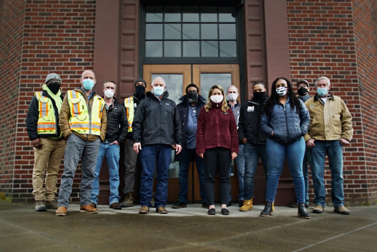 Grupo de 12 personas con máscaras de pie frente a un edificio posando para una foto grupal.
