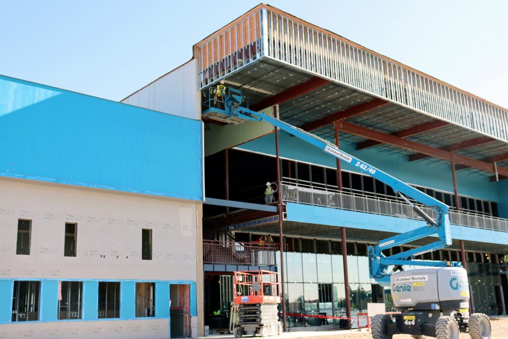 Drywallers on man lifts install metal corner beads on a glass facade of a 3 story building