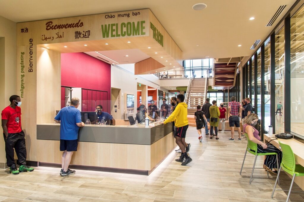 rec center patrons congregate at the front welcome desk with people using workspace and basketball courts in background