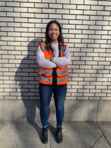 Una mujer con un chaleco naranja de construcción parada frente a una pared posando para una foto.