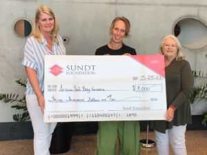 Three women holding a large check, standing inside the Sundt Tempe lobby.