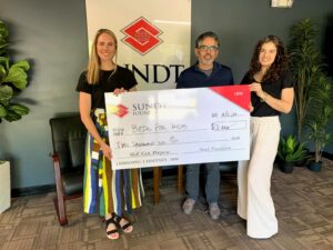 Three people holding a large check in front of Sundt office sign.