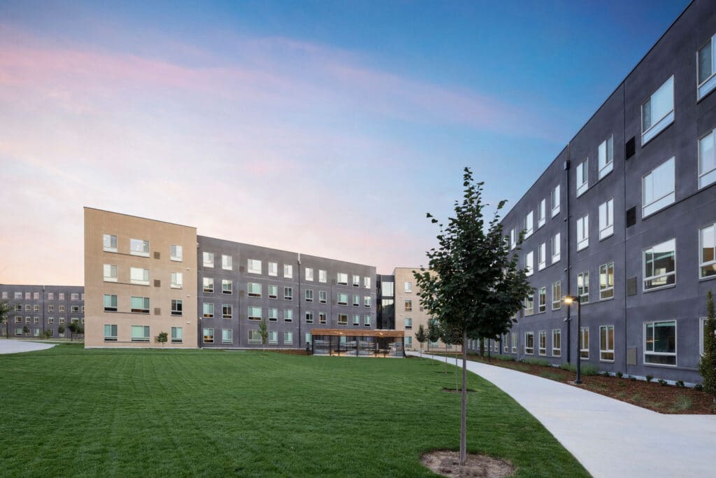 Multiple four-story buildings facing a grassy area and small trees lining a sidewalk.