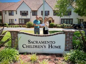 Two people holding a small check stand behind a sign that reads "Sacramento Children's Home". 
