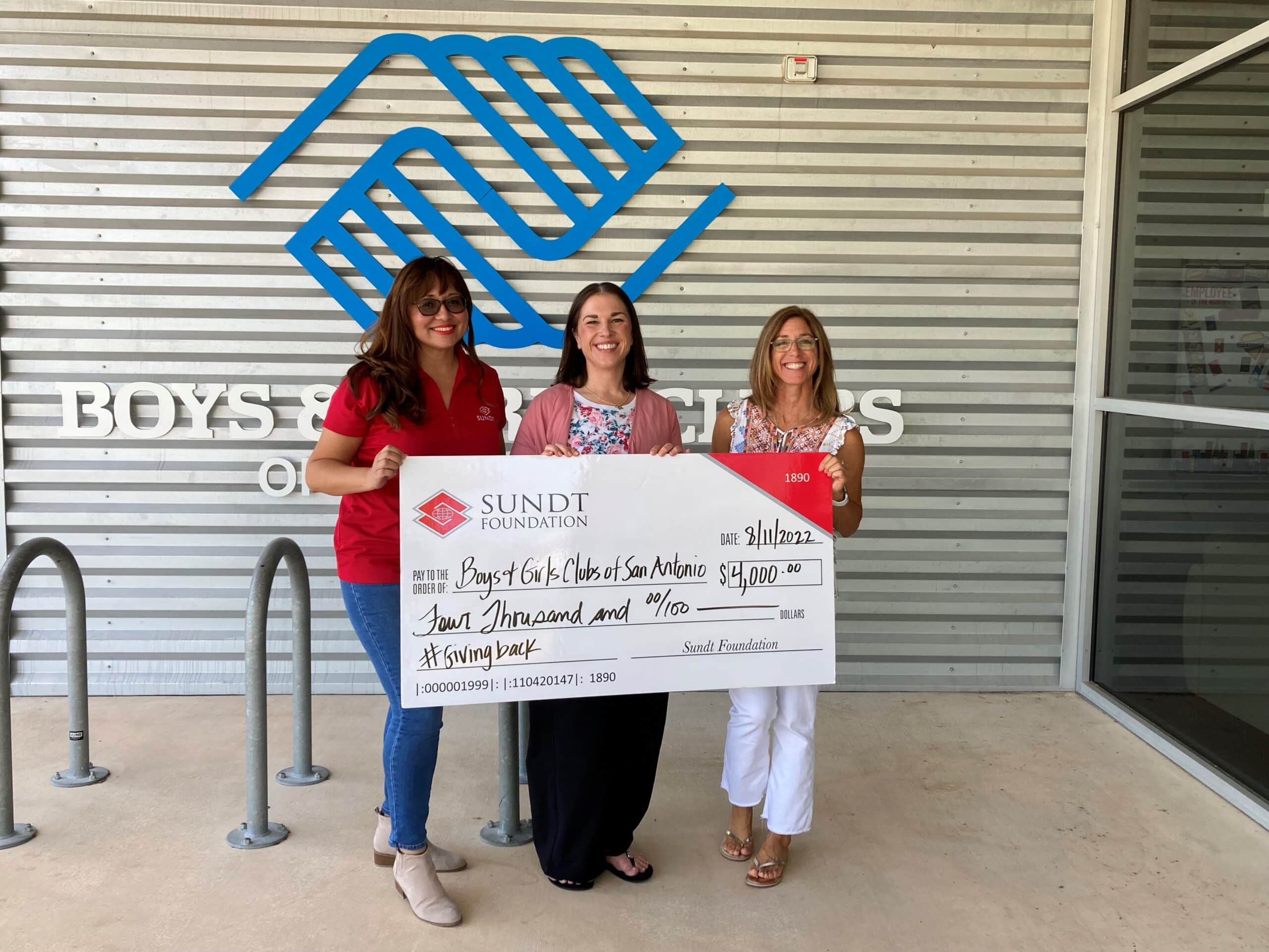 Three women standing in front of a wall with a charity logo and holding a check.